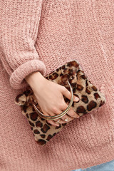 Close-up cropped shot of a girl, wearing pale pink knitted sweater. She's holding beige leopard print faux fur bag with metallic semicircular bag handles. There are two golden rings on her fingers. 
