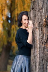 Beautiful elegant woman standing and posing near tree in a city park in autumn