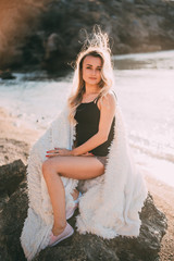 Beautiful girl with beautiful hair in shorts sitting on a rock on the background of rocks and the sea or ocean, wrapped in a fluffy light plaid