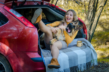 A happy Caucasian girl and a German shepherd lie in the trunk of a car in nature. Have fun, play and laugh. Friendship of man and dog, travel, camping on the autumn background. Moment of happiness
