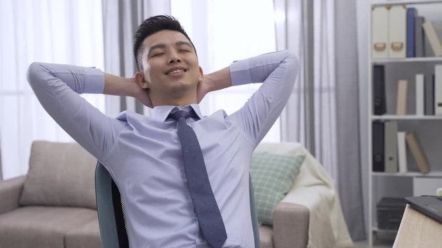 Cheerful young businessman sitting at work desk in home office and stretching body. happy asian korean man freelance worker with hands back on neck and head smiling daydreaming on chair in workplace