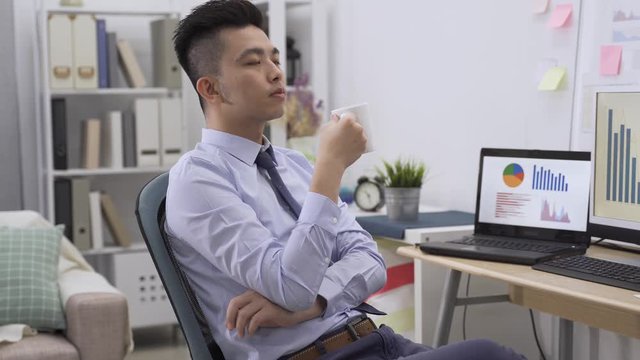 Finally coffee break. Tired young man in formal wear holding cup of tea and drinking it while sitting at workplace home office. exhausted male worker fell asleep while enjoy and relax with beverage
