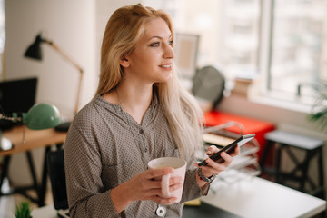 Close up of businesswoman in office drinking coffee and using phone.