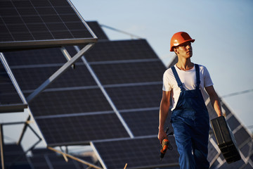 Cordless screwdriver in hand. Male worker in blue uniform outdoors with solar batteries at sunny day