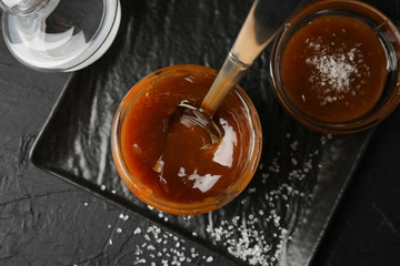 Glass jar with salted caramel and spoon on black background, top view