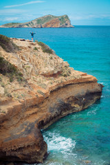 View of high cliffs above the turquoise sea line in Cala Escondida in Ibiza, Spain, white young male guy tourist waving and enjoying the view on top of a cliff