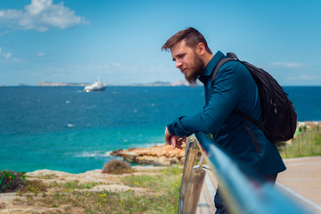 Handsome young white male tourist with a backpack leaning on a railing near a beach enjoying the view of green emerald water on Ibiza island