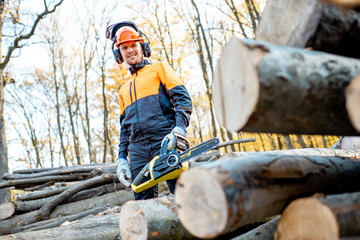 Portrait of a handsome professional lumberjack in protective workwear with a chainsaw near the wooden logs in the forest