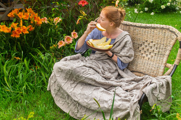 Red haired beautiful teenager girl in garden eating sliced melon, seating on a couch under beige...