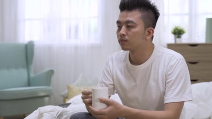 Young asian chinese man sitting in bed drinking water in mug. male enjoy breakfast beverage in early morning bedroom. attractive guy in tshirt relax and having cup of coffee to keep him awake at home - Powered by Adobe