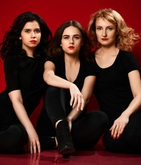 Closeup. Three young sexy women in black t-shirts and black tight jeans are sitting on the floor on red