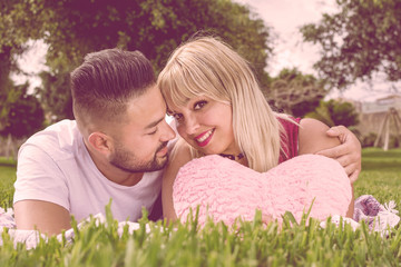 Portrait of a beautiful couple in love, lying on the grass with a pink heart-shaped cushion.