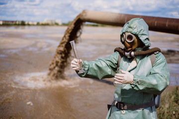 A male environmentalist in a protective suit and gas mask takes a sample of water. Scientist doing...