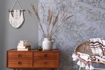 Stylish and boho composition of living room with design armchair, wooden retro commode, books, flowers in vase, beige macrame, plaid and elegant accessories. Modern home decor. Template. 