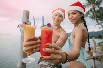 Young couple with cocktails in Christmas caps on the ocean on a tropical island.