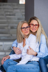 A beautiful blonde with glasses and her mature mother are sitting on the stairs outdoor. A well-groomed elderly woman hugs her adult daughter tightly. Love and family values. Mothers Day.