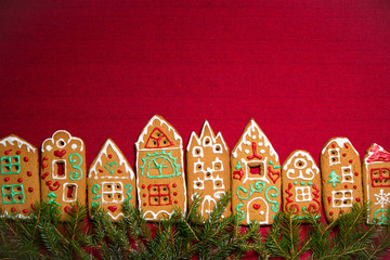 Christmas background. Red table cloth with town of cute gingerbread houses decorated with icing, Christmas lights, glitter and fir tree branches. Holiday mood. 