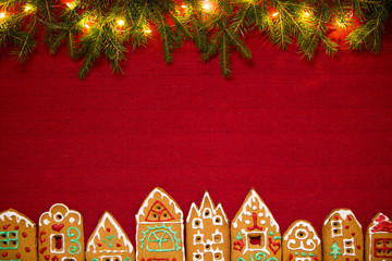 Christmas background. Red table cloth with town of cute gingerbread houses decorated with icing, Christmas lights, glitter and fir tree branches. Holiday mood. 