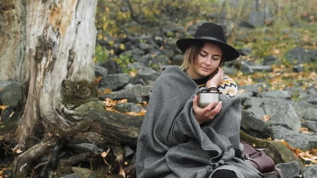 Portrait of beautiful woman in black hat sitting on rocks on cool autumn day near riverBank smiles and Drinks hot tea or coffee from a thermos. It is covered with a warm blanket and basks in nature.