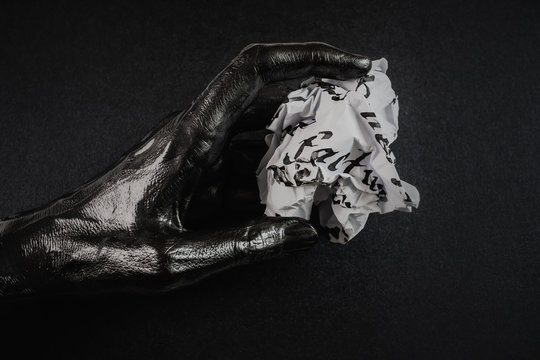 The hand is painted black with a metallic sheen. In hand is a crumpled sheet of paper with written ink and pen letters. Light background. Minimalism.