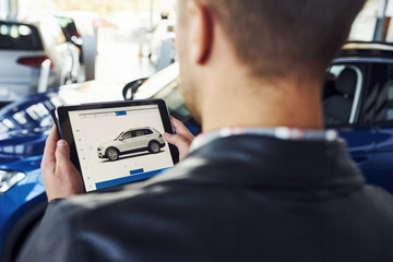 Man stands inside car salon with tablet in hands and looks at the vehicle picture