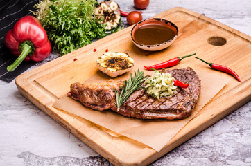 Medium rare grilled beef steak on a rustic cutting board with rosemary and spices, bbq sauce, red chili pepper, fried garlic, vegetables at a table restaurant background.