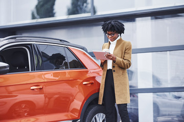 Young african american woman in glasses and with cup of drink stands outdoors near modern car