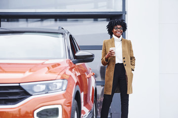 Young african american woman in glasses and with cup of drink stands outdoors near modern car