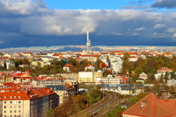 Panorama Of Prague. Houses with orange roofs, in the sunlight, among yellowed trees against a blue sky with stormy dark blue clouds, a TV tower in the distance, a railway.
