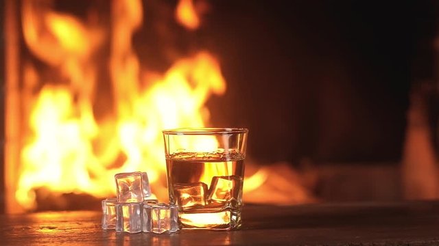 Slow-motion shot of Whiskey with ice for fun party against background of bright sparks and burning flames in fireplace. glass of old aged drink on the table with ice cubes in the middle of the frame.