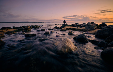 A person squats and looks out over the water by the sea with sunset in the background and rocks and a river in the foreground
