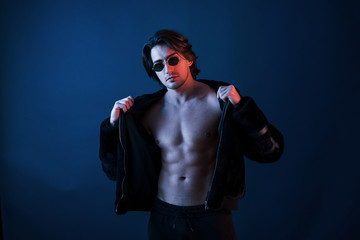 Fototapeta na wymiar Beautiful man in glasses and black clothes is in the studio with blue neon lighting