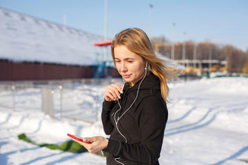 Portrait of a cute blonde girl listening to music while walking down the street holding a red phone in hand. Snow lies around