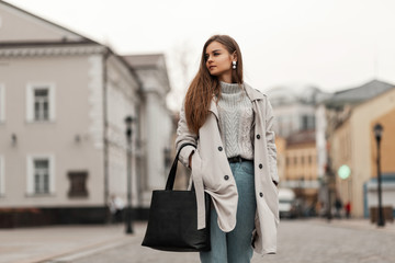 Charming young woman in a fashionable knitted sweater in a chic trench coat in blue jeans with a...