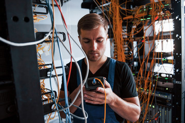 Young man in uniform with measuring device works with internet equipment and wires in server room
