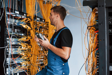 Young man in uniform with measuring device works with internet equipment and wires in server room - obrazy, fototapety, plakaty
