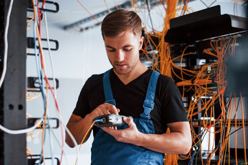 Young man in uniform with measuring device works with internet equipment and wires in server room