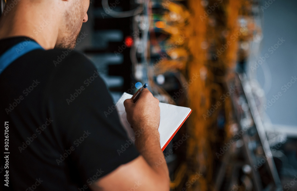 Wall mural young man in uniform with notepad in hands works with internet equipment and wires in server room
