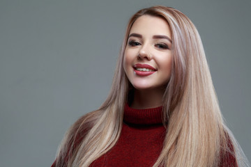 Beautiful smiling blonde girl with long hair in a warm burgundy sweater isolated on a gray background.