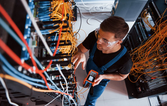Top view of young man in uniform with measuring device that works with internet equipment and wires in server room