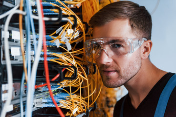 Young man in protective glasses works with internet equipment and wires in server room