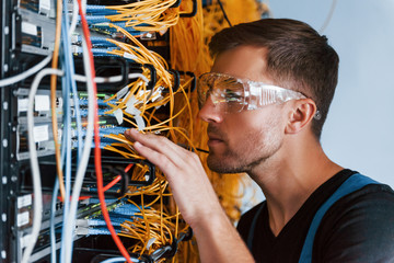 Young man in protective glasses works with internet equipment and wires in server room
