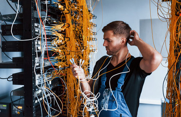 Young man in uniform feels confused and looking for a solution with internet equipment and wires in server room - obrazy, fototapety, plakaty