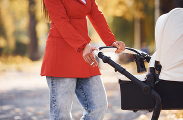 Close up view. Mother in red coat have a walk with her kid in the pram in the park at autumn time