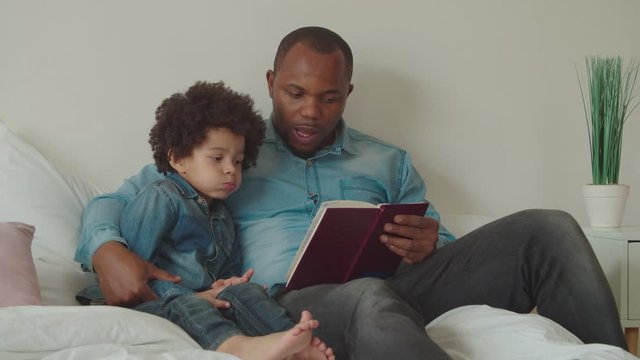 Caring loving black daddy embracing adorable preschooler multiethnic boy with curly hair, reading interesting fairy tale together on bed. Positive diverse family with child enjoying leisure at home.