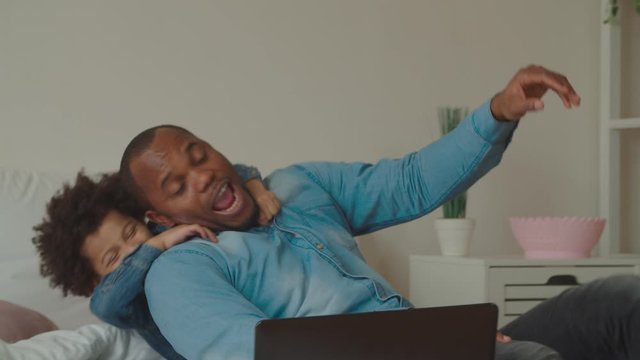 Playful excited little mixed race son embracing from behind loved joyful black father, having fun and joking while spending leisure together in bedroom, expressing happiness and carefree mood.