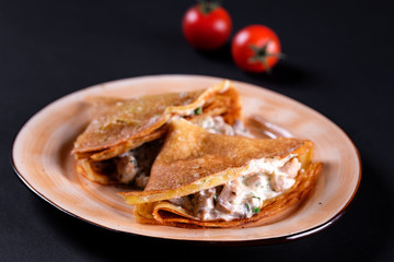 Delicious crepes pancake with champignon cream, chicken, mushrooms fresh green herbs and white cheese on plate, black background. Top view homemade breakfast. Russian national food.