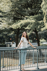 young woman is happy, lady is smiling, woman in white t shirt has happiness, asian girl closing eyes and smiling with hands on railing, chilling moment at the bridge and trees background