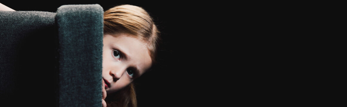 panoramic shot of scared child looking at camera while hiding behind armchair isolated on black
