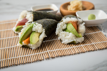 Lachs Fisch, Tunfisch und Surimi Avocado Temaki Sushi, eingelegter Ingwer mit Soja Soße und Sushimatte auf Marmor Hintergrund